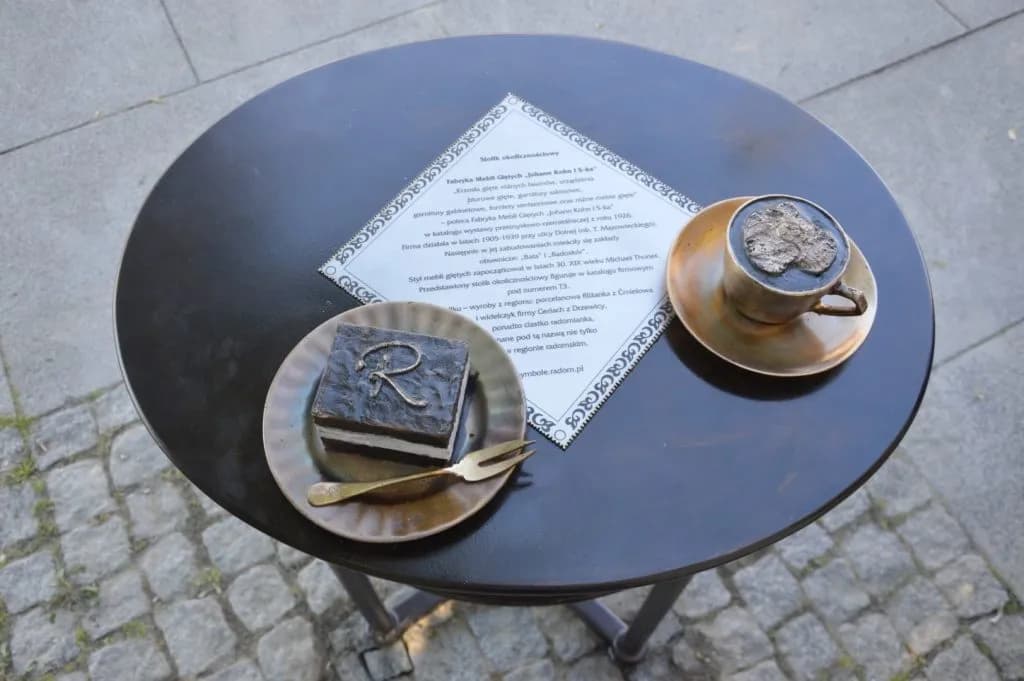 A bronze sculpture depicting a slice of cake with a fork and a coffee cup on a black table with a diamond-shaped informational plaque.